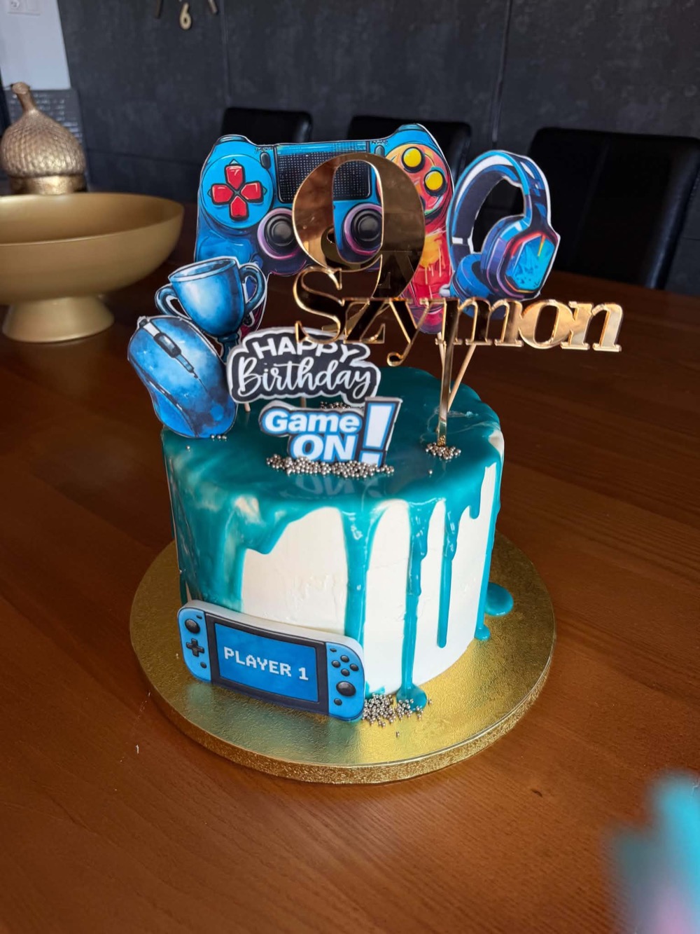 Turkusowy tort urodzinowy typu drip cake z gamingowymi toperami: padem, słuchawkami i napisem Game ON